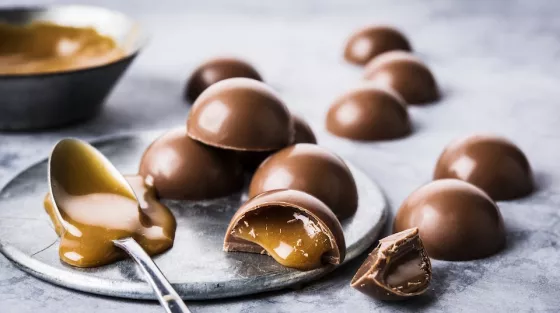 Round bonbons on a plate and marble tabletop with a bowl of caramel sauce in background, spoon of spilling caramel sauce in spoon and coming from cut bonbon